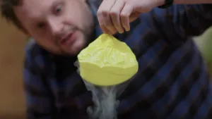 Een foto van presentator Diederik Jekel die een samen getrokken ballon vasthoudt boven een bak met een heel koude vloeistof. Er is damp te zien aan de onderkant van de ballon.