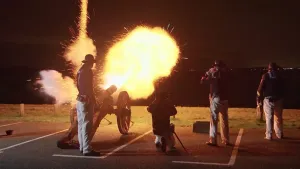 Een foto van een oude kanon die in het donker wordt afgeschoten. Er komt een grote vlam uit het kanon die alles verlicht. Er staat een man vlak achter het kanon en er staan nog meer mannen naast hem.