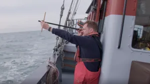 Een foto van Diederik Jekel op een schip. Hij heeft een graadstok in een uitgestrekte hand vast en houdt het andere eind van de stok bij zijn oog. Zo kijkt hij schuin omhoog. Op de achtergrond zie je de zee.