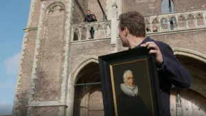 Een foto van een man die een schilderij van het gezicht van een man voor de camera vasthoudt. Hij kijkt naar presentator Jekel die achter hem staat op een balkon van een oude toren.