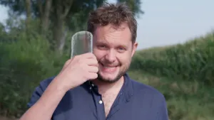 Een foto van presentator Diederik die buiten op een weg loopt met de camera vlak voor hem. Hij laat een stuk glas zien die hij naast zijn gezicht houdt.