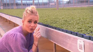 Een foto van presentator Janouk die op de grond zit van een kweekkas. Naast haar staat er een grote bak waar een heleboel kleine potjes met plantjes in staan, in de foto zijn er honderden te zien..