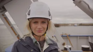 Een foto van presentator Janouk aan boord van een schip. Ze kijkt recht in de camera en heeft een grote witte helm op. 