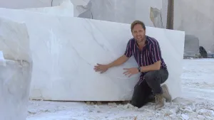 Een foto van een presentator die op zijn knie zit en zijn rechterhand heeft liggen op een groot, rechthoekig blok marmer dat wit is, met grijze kronkelige lijnen in het wit. Marmer is een steensoort ontstaan in de Italiaanse bergen uit kalk. 