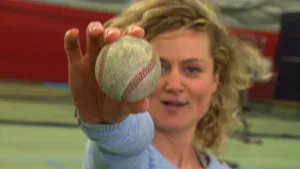 Een foto van een presentator in een gymzaal met groene vloer, die een honkbal omhoog houdt naar de camera toe. De honkbal is wit, maar zwarter geworden door het slaan met een honkbalknuppel en er lopen rode draadjes door de bal. 