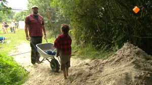 Een foto van presentator boswachter Tim die naast bomen en struiken tegenover een kind staat met een kruiwagen. Achter het kind is een grote hoop met zand. Achter boswachter Tim is er een grasveld te zien waar nog meer mensen op staan.