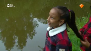 Een foto van een meisje die aan de oever staat van het water en naar boven kijkt. Achter haar staat er nog iemand die haar haar vasthoudt.