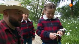 Een foto van boswachter Tim die naast een meisje zit die met haar handen bezig is met een witte bloem. Achter hun staat een jongetje te kijken en zijn er nog meer van dezelfde witte bloemen te zien.
