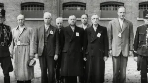 Een foto in zwartwit van ongeveer tien mannen die naast elkaar staan. Ze kijken richting de camera en ze kijken niet blij. Op hun kleren zit een kaartje vastgemaakt. Op de achtergrond zie je een gebouw dat een gevangenis zou kunnen zijn.