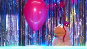 Een foto van een pop die Skip de Plofkip heet. De pop heeft een wit verenpak, een gele snavel en een rode kam op haar hoofd. Met ronde, bolle ogen kijkt ze naar een roze ballon links in beeld.  Erachter hangen glinsterende, zilverkleurige strookjes.