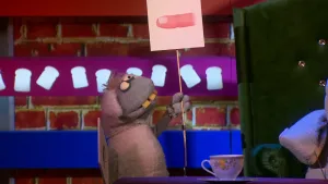 Een foto van pop Stella Schaap met een witte vacht die grijs is bij kop en poten, met ronde ogen en vooruitstekende, gele tanden. De pop heeft een bord met een getekende vinger erop vast en kijkt ernaar. Rechts is de groene stoel van Nico te zien.