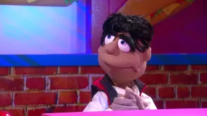 Een foto van een pop die Thomas Achterdocht heet achter een roze tafel. Hij heeft zwart haar, grote ogen met blauwe pupil en dikke, zwarte wimpers. Hij draagt een rood, mouwloos jasje. Achteraan hangt een tekening van een broodje kaas, sla en tomaat.