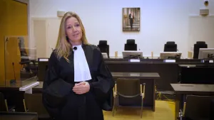 Een foto van de officier van justitie in een zwart pak met brede mouwen en een witte, rechthoekige doek om haar nek. Ze heeft haar handen over elkaar heen gevouwen. In de rechtzaal erachter staan stoelen en tafels voor rechters, advocaten en de verdachte.