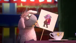 Een foto van pop Stella Schaap. De pop heeft een bord vast met een getekende, oude man erop, met een zwarte bril en een grijze snor en haar. De pop heeft een witte vacht die grijs is op haar kop en poot, ronde ogen en vooruitstekende, gele tanden.