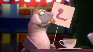 Een foto van pop Stella Schaap. De pop heeft een bord met een getekende roze worm erop vast en kijkt naar het bord. De pop heeft een witte vacht op haar rug en oren, een grijze vacht bij kop en poten, ronde ogen en vooruitstekende, gele tanden.