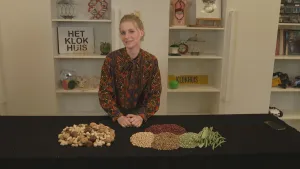 Een foto van presentator Janouk. Ze staat achter een tafel in de tv-studio. Op die tafel liggen allemaal noten en zaden, netjes gesorteerd. Op de achtergrond zie je spullen van het Klokhuis in open kasten.