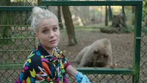 Een foto van presentator Janouk. Ze staat voor een metalen hek in een bos en kijkt in de camera. Achter dat hek zit een bruine beer. Hij kijkt naar iets op de grond. 