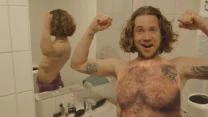 Een foto van presentator Pascal die in de badkamer in de camera kijkt en zijn spierballen laat zien. Er is een foto geplaatst van een borst vol met borsthaar bovenop zijn echte borst.