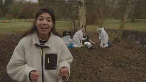 Een foto van presentator Manon. Ze vertelt wat in de camera. Op de achtergrond zie je twee mannen in witte pakken. Ze zitten geknield bij een persoon of een pop, die tegen een boom aan ligt. Op de achtergrond zie je een rood-wit gestreept lint.