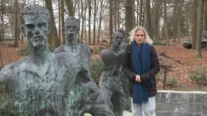 Een foto van presentator Janouk. Ze staat in een bos of een park en kijkt in de camera. Naast haar staan enkele bronzen beelden van mannen. De beelden kijken ongelukkig. In de achtergrond zie je een tank staan. 
