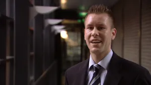 Een foto van een man die een zwart pak met een lichtpaarse blouse eronder aanheeft en een zwarte stropdas met grijze, witte en gele strepen erop om zijn nek. Hij staat in een lange gang waar rechts dichte lichtbruine rolluiken te zien zijn. 