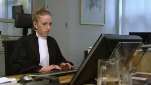 Een foto van iemand die werkt als griffier in een zwart kostuum en met een rechthoekige, witte lap om de nek. De griffier tikt op een toetsenbord en kijkt naar een beeldscherm. Er staan ook een muis, vaste telefoon en een kan water met glas op het bureau.