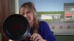 Een foto van een presentator die een koekenpan voor haar gezicht houdt en naar de achterkant van de pan kijkt. 
