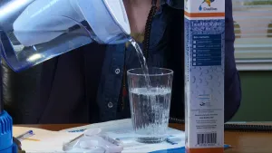 Een foto van iemand die water in een glas giet uit een kan waar een filter in zit waar het water doorheen. Naast het glas staat er een lange doos rechtop op tafel waar een water filter in zit.