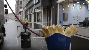 Een foto van een snackbar vanaf de straat gezien. Op de voorgrond zie je een groot beeld van een zak met friet, als reclame. In de snackbar zie je een man.