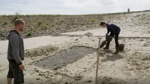 Een foto van twee mannen die in het zand staan. Een van hen gooit een emmer met zand of iets dergelijks leeg. De ander kijkt toe. Op de achtergrond zie je een heuvel, met stenen daarop. 