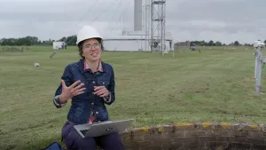 Een foto van een vrouw met een bouwhelm op. Ze zit op een stenen muurtje en heeft een laptop op schoot. Ze praat tegen de kijker. Op de achtergrond zie je apparaten voor weerkundigen.
