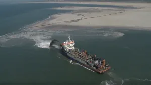 Een foto vanuit de lucht genomen. Je ziet de zee met daarin een groot schip. Dat schip spuit zand in zee om zo een stuk land te maken. Daarachter zie je al een droog strand dat gemaakt is. 