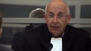 Een foto van een rechter die van dichtbij te zien is. De rechter heeft een zwart kostuum dat voor een rechter is aan en een wit, rechthoekig lapje om zijn nek hangen. Vóór de rechter is een microfoon met een zwart bolletje erop te zien.