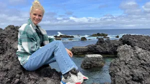 Een foto van presentator Janouk die aan de kust op de rotsen zit. Op de achtergrond is de zee en nog meer rotsen te zien.