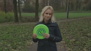 Een foto van presentator Janouk die buiten in een park een frisbee vasthoudt terwijl ze de camera inkijkt.