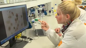 Een foto van Janouk in een witte laboratoriumjas en iemand in dezelfde jas, waarvan het gezicht niet te zien is. Ze wijst naar een beeldscherm op tafel, met bruine, ronde vlekken en vlekjes erop. Dat zijn tumorcellen van mensen met de ziekte kanker.