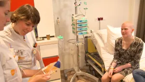 Een foto van Emma die kaal is en op een bed zit. Links staan twee verpleegsters of dokters. Eén kijkt in een doorzichtig plastic zak met vloeistof die de ander vastheeft. Het is de chemokuur die Emma krijgt met een infuus, een buisje met een naald.