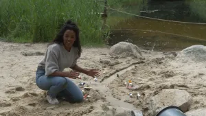 Een foto van Sosha die op haar knie zit op een zandvlakte. Ze wijst met beide handen naar een uitgegraven muur van zand, een dijk, die langs een slingerende rivier van zand naar het water achter Sosha loopt. Er staan plastic poppetjes en dieren naast.
