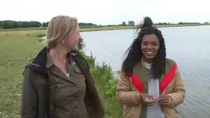 Een foto van Sosha en boswachter Lianne op een groen grasveld langs de waterkant, een geul langs de rivier de Maas. Lianne draagt een donkergroene blouse en jas en Sosha een lichtbruine jas met schuine, rode en groene streep en een lichtgrijze trui.