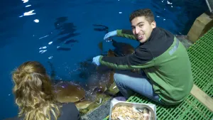 Een foto van presentator Nizar die aan het water zit van een aquarium en haaien aan het voeren is met vis. Naast hem zit er een vrouw die toekijkt.