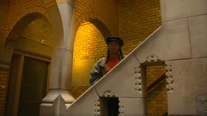 Een foto van Eva in de Burcht van Berlage, achter een lichtbruine stenen leuning op een trap. Linksboven haar is een boog van rode en lichtbruine stenen te zien. Door een opening in de stenen leuning, is de houten trapleuning aan de muur te zien.