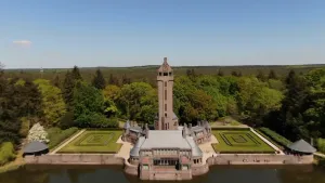 Een foto van een stenen gebouw, Jachthuis Sint Hubertus. Vooraan ligt water achter een muur met aan beide kanten ronde, grijze daken. Het gebouw heeft een grijs dak, een grote toren met schuin dak achteraan en een tuin van groen gras aan beide kanten. 