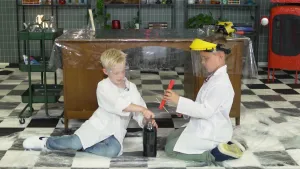 Een foto van Calvin en Sybren in een witte jas en plastic beschermmasker. Ze zitten op een doorzichtige, plastic beschermlaag op de vloer. Sybren doet een plastic fles met cola open, Calvin heeft een buis van rood karton vast met mentossnoepjes erin.