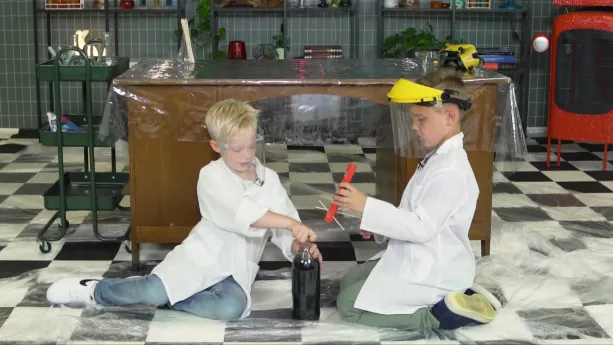 Een foto van Calvin en Sybren in een witte jas en plastic beschermmasker. Ze zitten op een doorzichtige, plastic beschermlaag op de vloer. Sybren doet een plastic fles met cola open, Calvin heeft een buis van rood karton vast met mentossnoepjes erin.