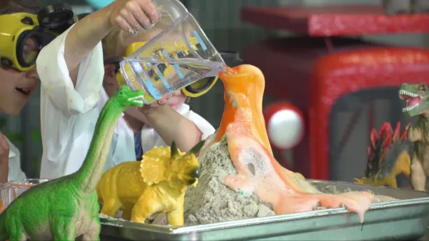 Een foto van Tijn en Sophie in een witte jas. Op tafel staat een zilverkleurige bak met een soort vulkaan erin: een zandkleurige hoop met roodkleurige vloeistof erop. Sophie giet er water op uit een doorzichtige kan. Er staan vier plastic dino's naast.
