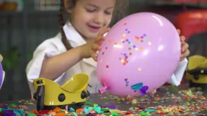 Een foto van Alyssa die een witte jas aanheeft en achter een tafel staat. Ze heeft een roze ballon vast, waar confetti opzit. De zwarte tafel ligt ook vol met confetti. Op de tafel staan ook twee geelbruine veiligheidsbrillen. 