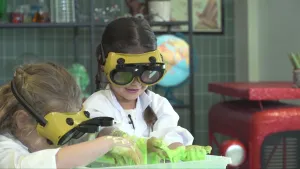 Een foto van Siem en Evelien. Hun handen hangen net boven een plastic bak en zitten onder het slijm. Alleen de bovenrand van de plastic bak is te zien. Ze dragen een witte jas en een bruingele veiligheidsbril met een zwarte, opengeklapte oogklep ervoor.