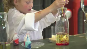 Een foto van Lynn die in een witte jas achter een tafel staat. Op de tafel staan drie flesjes met rode, oranje en blauwe vloeistof, olie, en een glazen vaas met rode olie op de bodem en gele olie erop. Lynn knijpt in een flesje gele olie boven de vaas.
