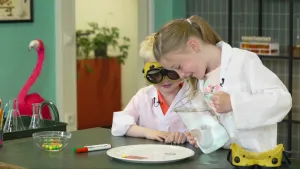 Een foto van Fox en Demi-Lynn achter een tafel met een wit bord, twee watervaste stiften en een schaal met ronde snoepjes erop. Demi-Lynn giet water uit een glazen kan op het bord met één geel, rond snoepje en twee niet goed zichtbare tekeningen erop.