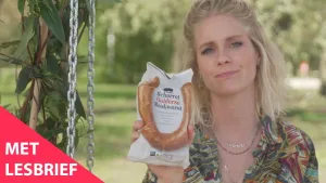 Een foto van presentator Janouk. Ze zit buiten en kijkt in de camera. Ze toont de kijker een voorverpakte rookworst. Linksonder in beeld staat de tekst "Met lesbrief".